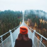 woman wearing knit cap walking on white bridge between trees during daytime